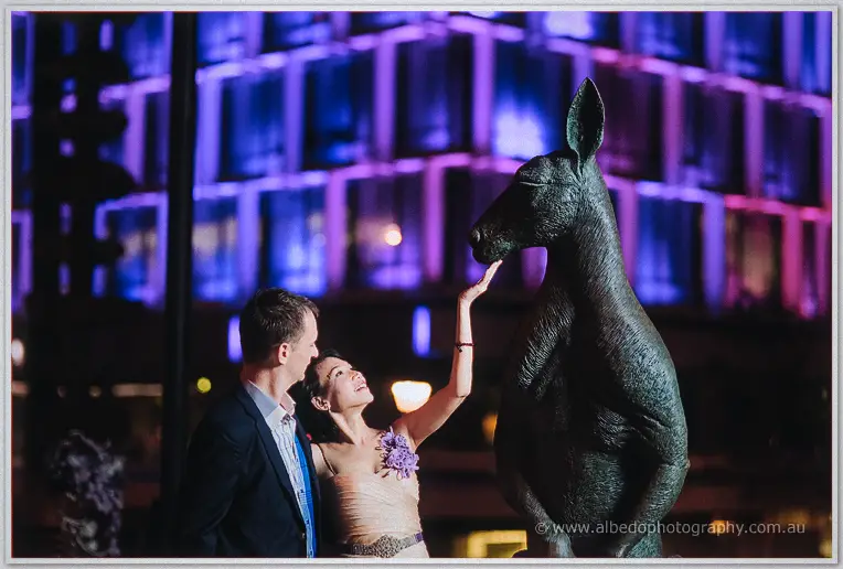 Loan beams as she reaches up to touch a bronze kangaroo, Alex beside her under Perth's purple night lights.