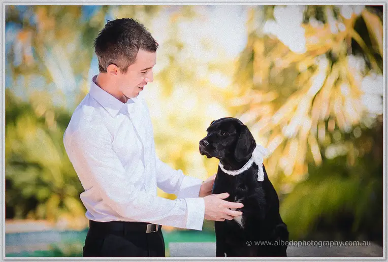 Groom in white shirt steadies a black dog wearing a white lace collar in golden garden light