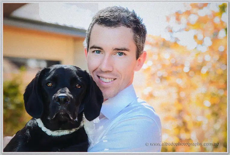 Groom cuddles his black labrador close to his shoulder in warm autumn backyard light
