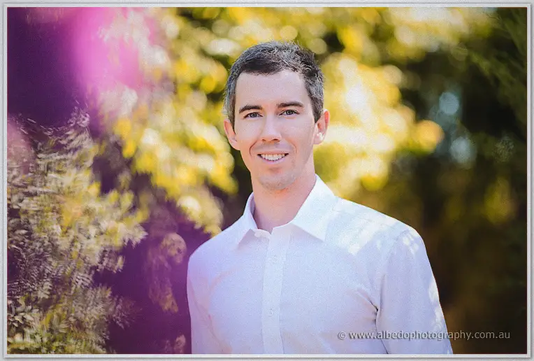 Groom smiles straight at the camera in a white shirt against sunlit foliage and purple flare