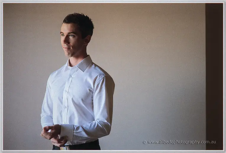 Thoughtful groom buttons his cuff against a plain taupe wall in soft window light