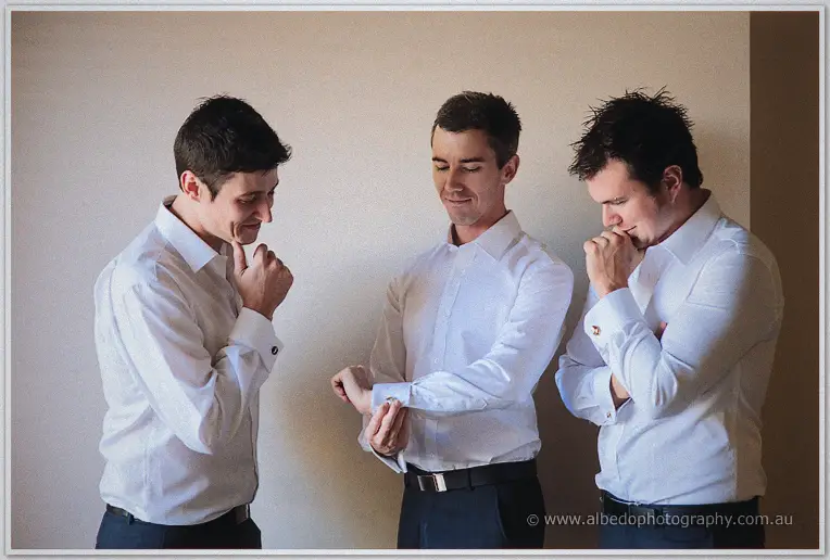 Groom fastens his cufflink while two groomsmen watch and smile during getting-ready moments