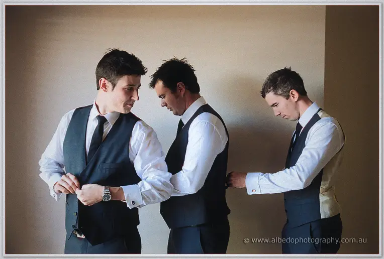 Three groomsmen in navy vests and white shirts help each other dress against a plain wall