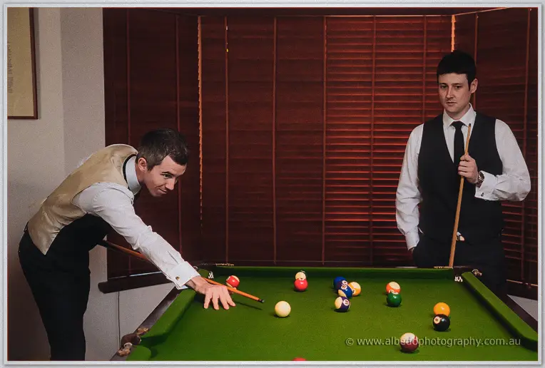 Groom lines up a pool shot while a groomsman watches with cue in hand by timber shutters