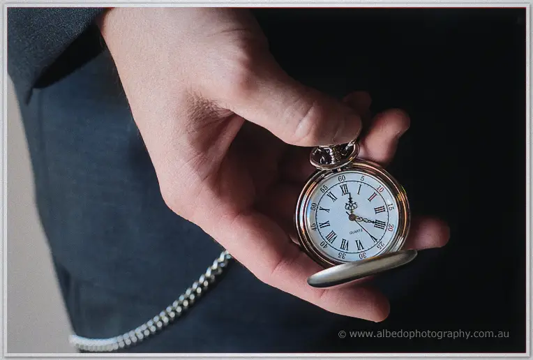Groom's hand opens a silver pocket watch with roman numerals against his dark suit