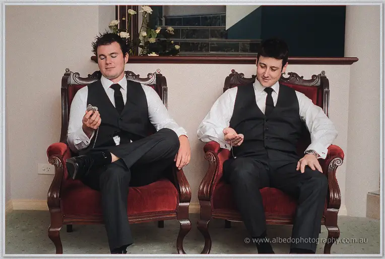Two groomsmen check pocket watches while seated in carved red-velvet armchairs indoors