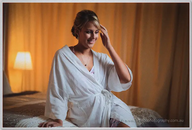 Lauren smiles softly in a white robe on the bed's edge amid warm lamplight