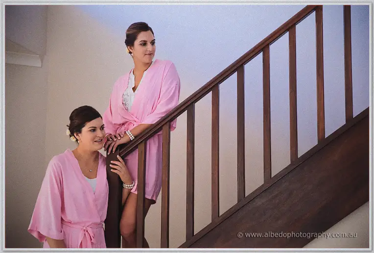 Bridesmaids in pink robes chat on a timber staircase with pearl bracelets and upswept hair