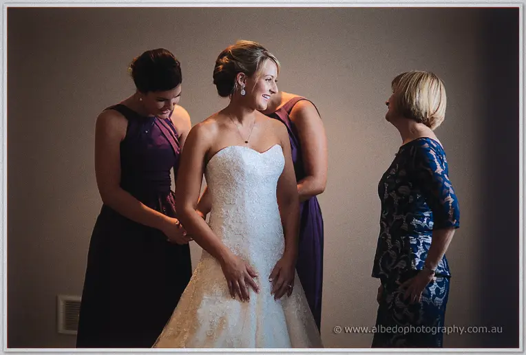 Bride Lauren smiling as bridesmaids in purple gowns and her mother help fasten her lace dress