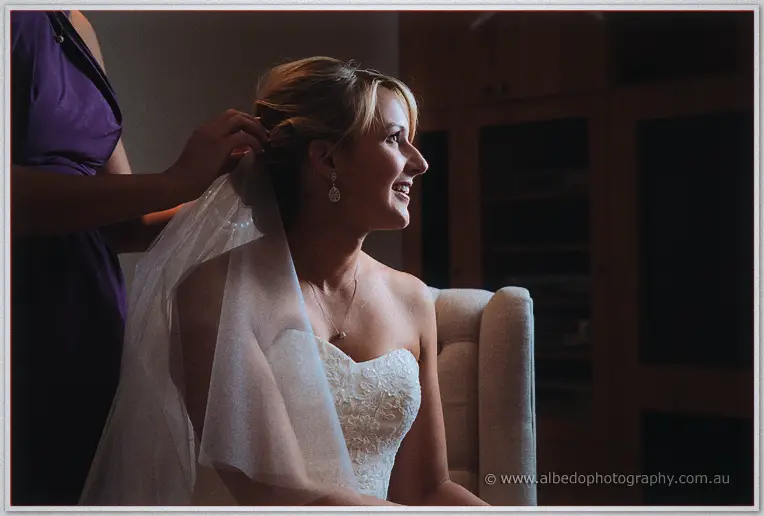 A bridesmaid pins Lauren's veil in place as she sits smiling in profile, warm low light