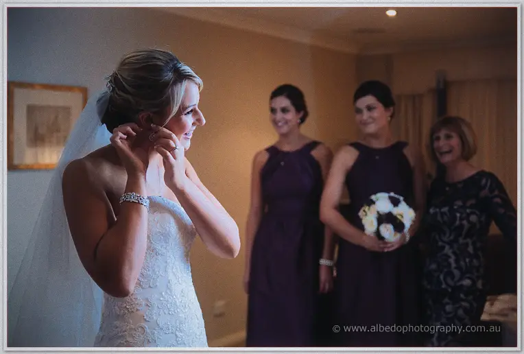 Lauren fastens an earring, laughing, as bridesmaids and her mother watch holding white and purple bouquets