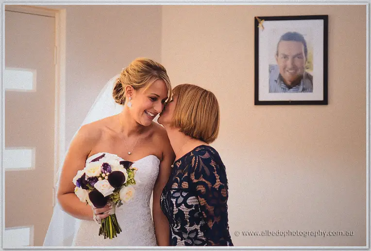 Lauren's mother kisses her cheek as she holds her bouquet, framed portrait glowing above