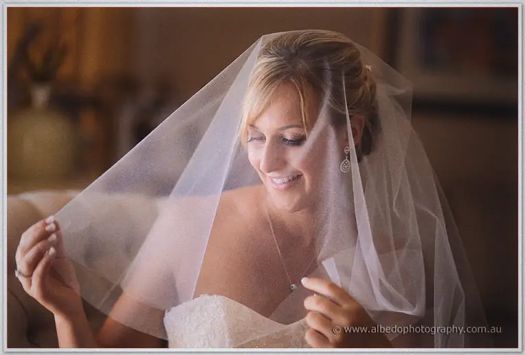 Lauren lifts her sheer veil over her face and smiles, eyes lowered in soft light