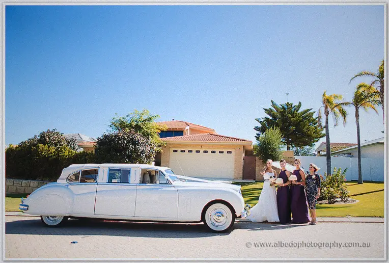 Lauren and her bridal party wait beside a white vintage limousine under blue Perth skies