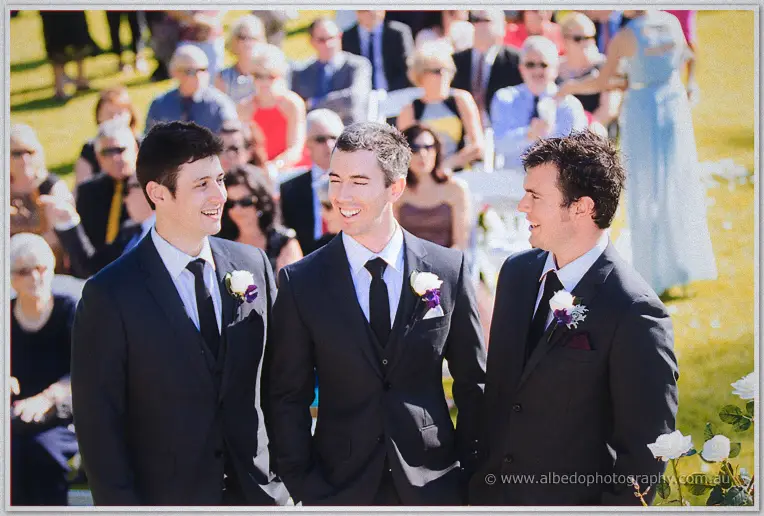 Groom Ryan grins with his groomsmen in charcoal suits before guests seated on the lawn