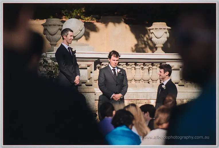 Ryan waits with his groomsmen beneath stone urns, guests' silhouettes in the foreground