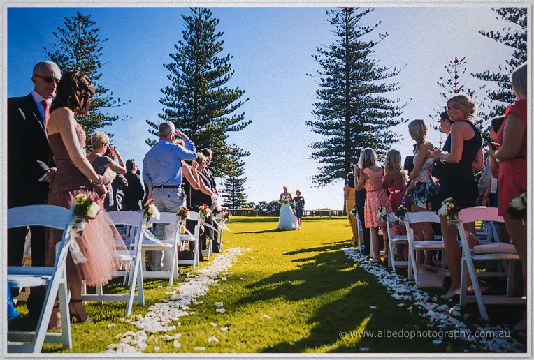 Lauren walks the petal-strewn aisle between white chairs under tall pines by the sea
