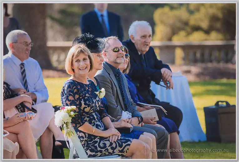 Wedding guests in spring dress watch the ceremony from white chairs in the gardens