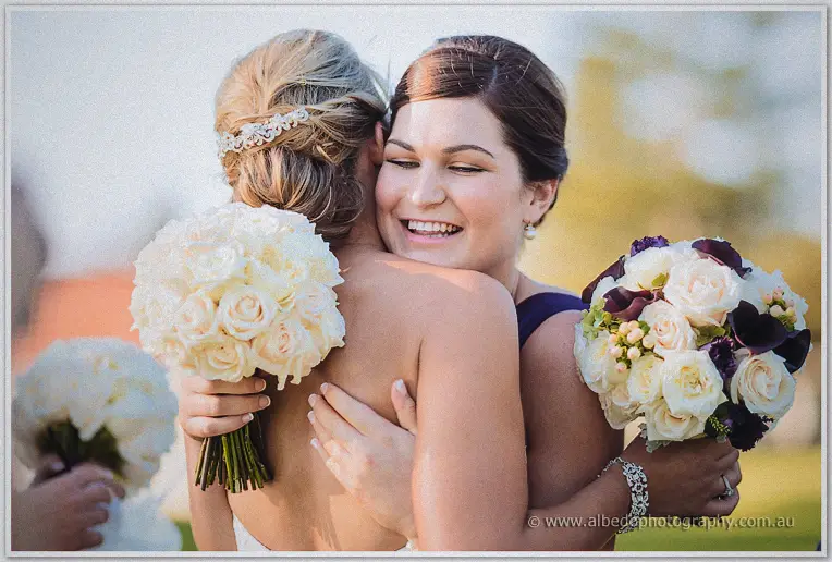 A bridesmaid hugs Lauren tightly, both grinning with bouquets in hand