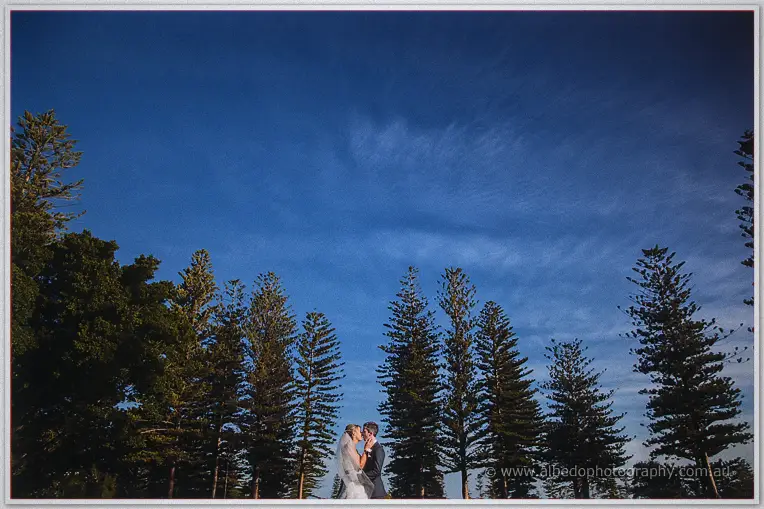 The newlyweds kiss on the lawn beneath Norfolk pines and a vast evening sky