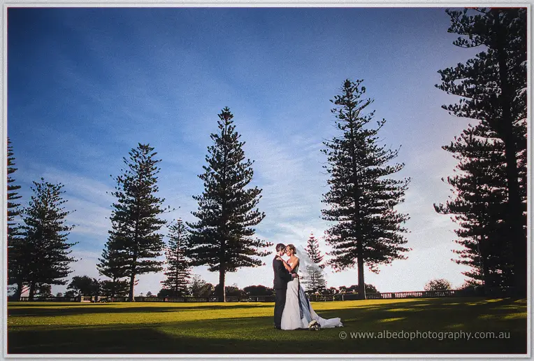 Lauren and Ryan embrace on the Cottesloe Civic Centre lawns beneath tall pines and a deep blue sky
