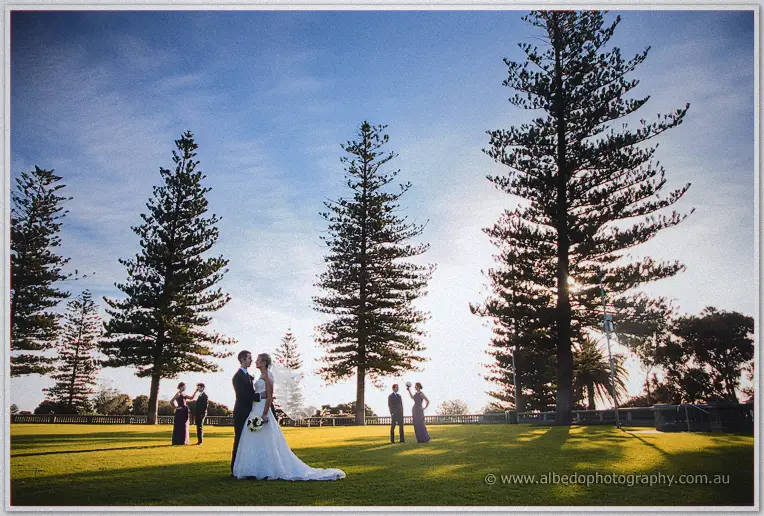 Lauren and Ryan pose on the lawn as bridesmaids in purple stroll beneath Cottesloe's towering pines