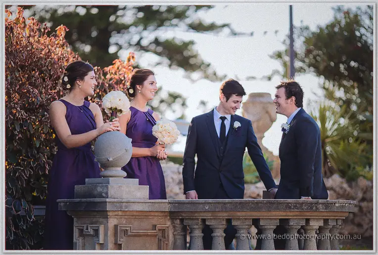 Bridesmaids in purple gowns laugh with groomsmen in navy suits by a stone balustrade in golden light