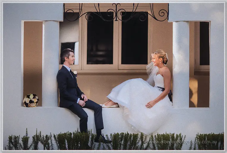 Lauren and Ryan sit facing each other on a white veranda ledge, laughing in afternoon light