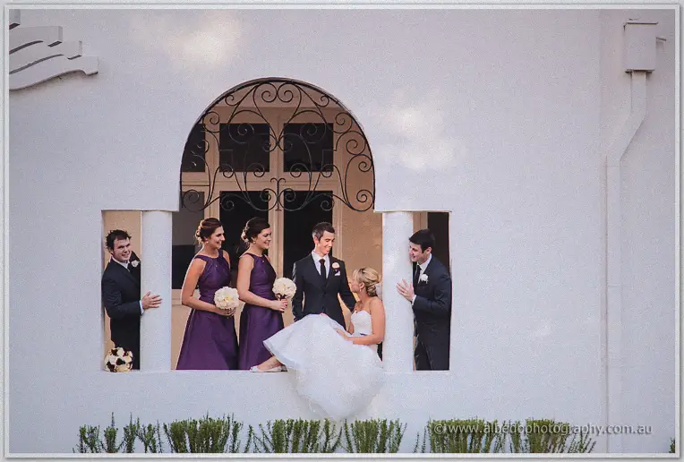 Bridesmaids in purple and groomsmen in navy crowd a Cottesloe archway around the seated couple