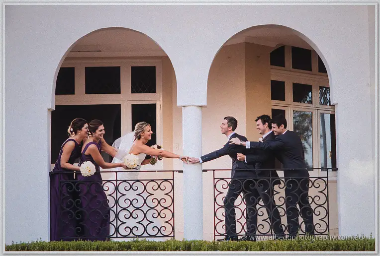 Lauren reaches playfully toward Ryan across twin veranda arches as the wedding party laughs