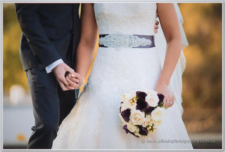 Detail of Lauren's jewelled sash, cream and plum rose bouquet, and hands entwined with Ryan's