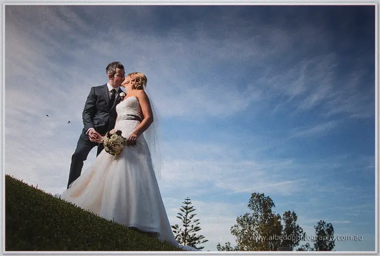 Lauren and Ryan kiss on a grassy rise under dramatic clouds streaked across the afternoon sky