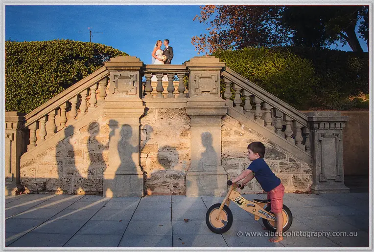 Lauren and Ryan pose atop a grand stone staircase as a boy rides his balance bike below