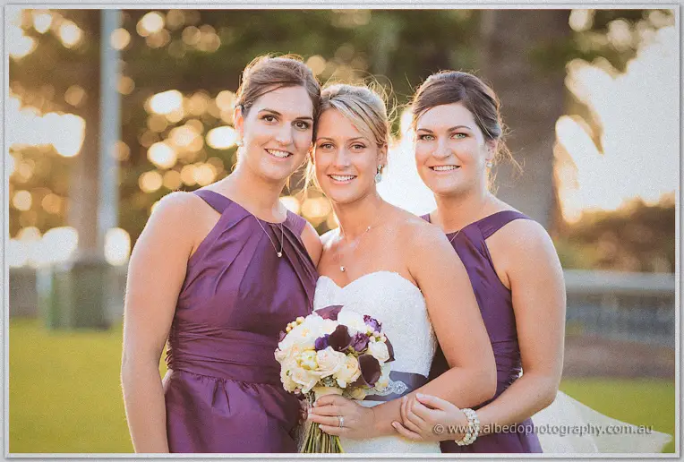 Lauren beams between her two bridesmaids in purple as sunset light glows through the trees