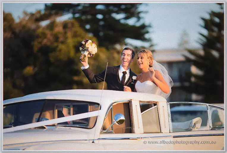Lauren and Ryan wave from the open roof of a white vintage limousine, bouquet raised in triumph