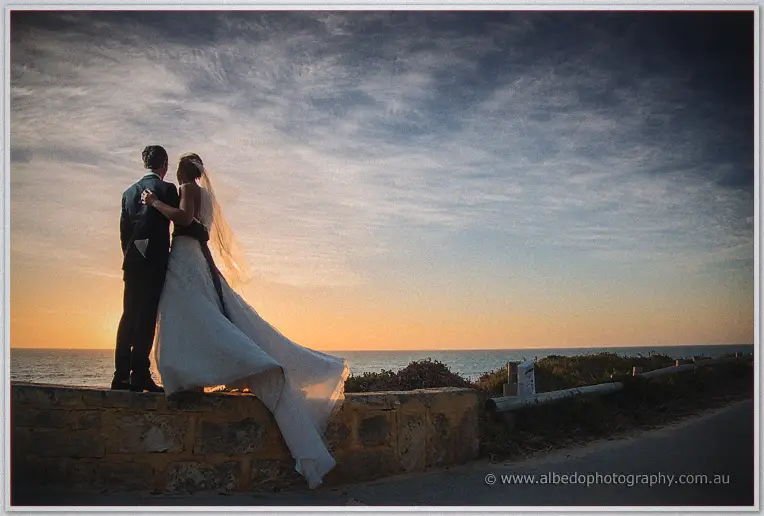 Lauren and Ryan stand on a limestone sea wall, her train streaming as the sun melts into the ocean