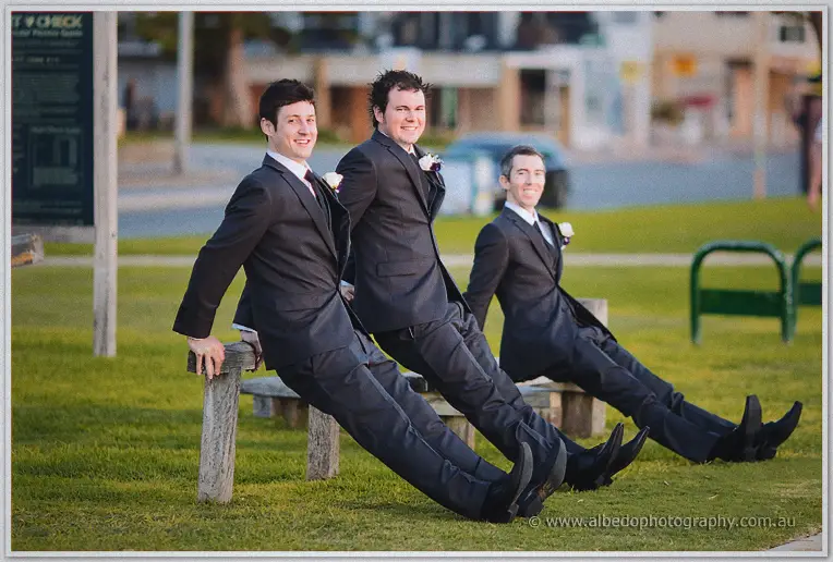 Groomsmen in suits lean back in a row on a foreshore bench, boots stretched toward the camera