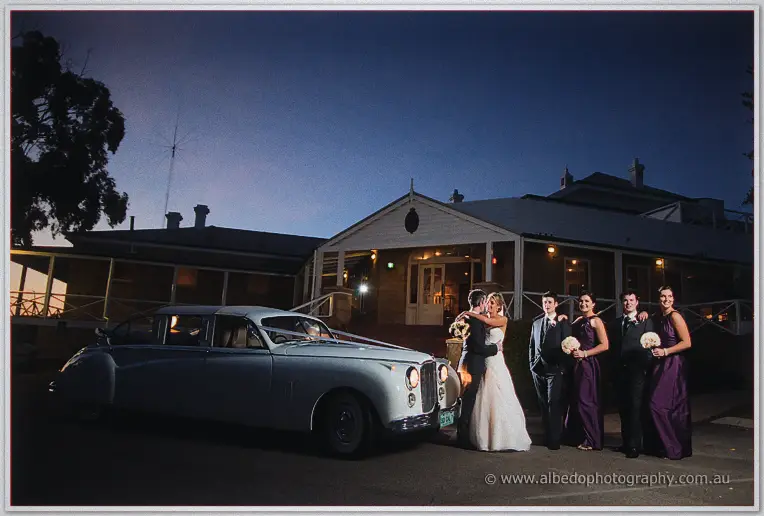 The couple and wedding party gather by a pale vintage Jaguar outside the yacht club at nightfall