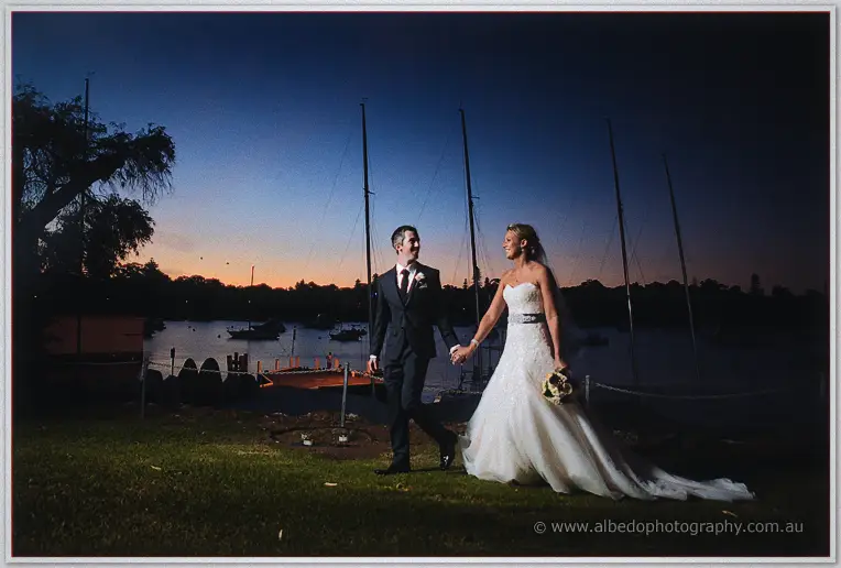 The couple stroll hand in hand along the riverbank at last light, yachts moored behind them