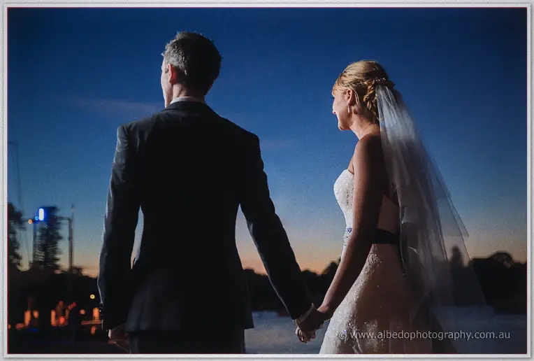 Seen from behind, the couple hold hands facing the deep-blue evening sky over the bay