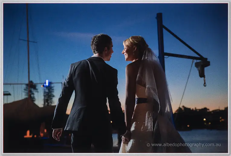 The couple glance back at each other on the dock, sailboats and rigging dark against dusk
