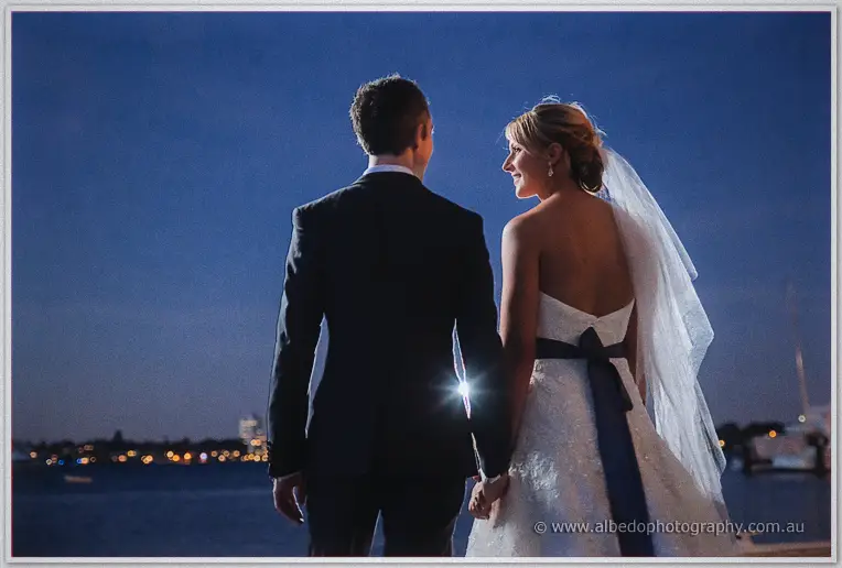 Bride and groom hold hands gazing across twilight bay waters in a Freshwater Bay Yacht Club portrait