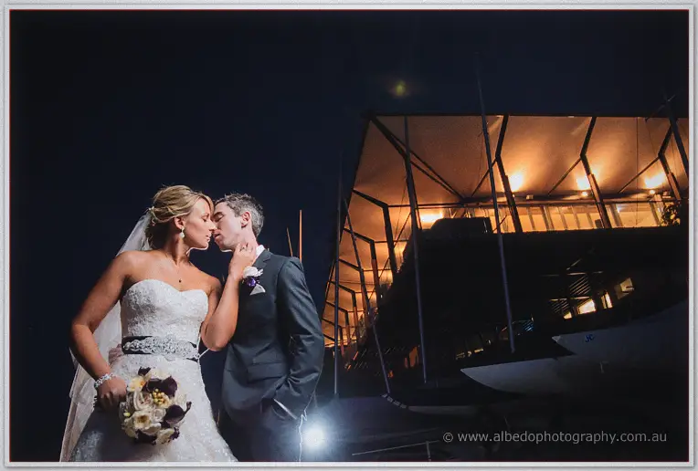 Lauren holds Ryan's face as they kiss in the marina night, clubroom lights glowing above