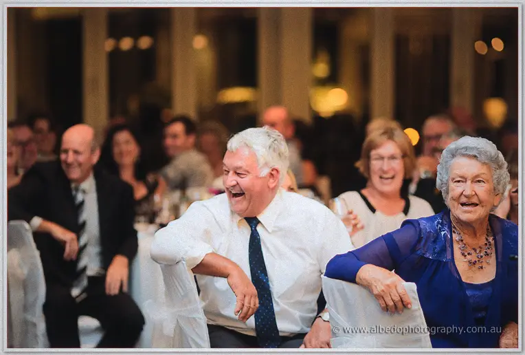 Guests double over laughing in the lamplit reception room, an older woman beaming in blue