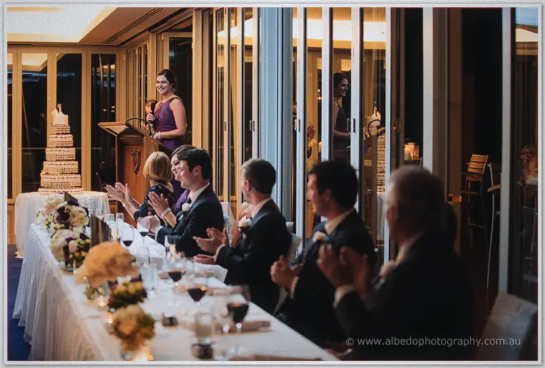 Bridesmaid in purple giving a speech at the lectern while the applauding bridal table sits beside a tiered cake
