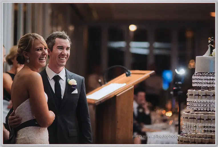 Lauren and Ryan laughing during speeches beside their tall tiered cake in the evening reception room