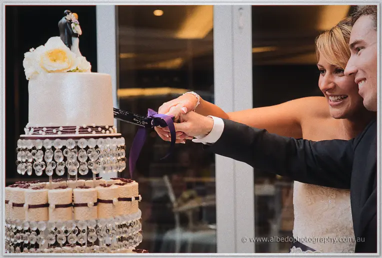 Lauren and Ryan cutting their crystal-hung tiered cake together, both beaming, hands joined on the knife