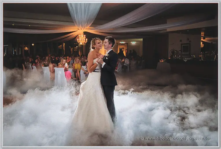 Wide view of the newlyweds dancing hip to hip, deep fog swirling around their gown and suit under draped ceilings