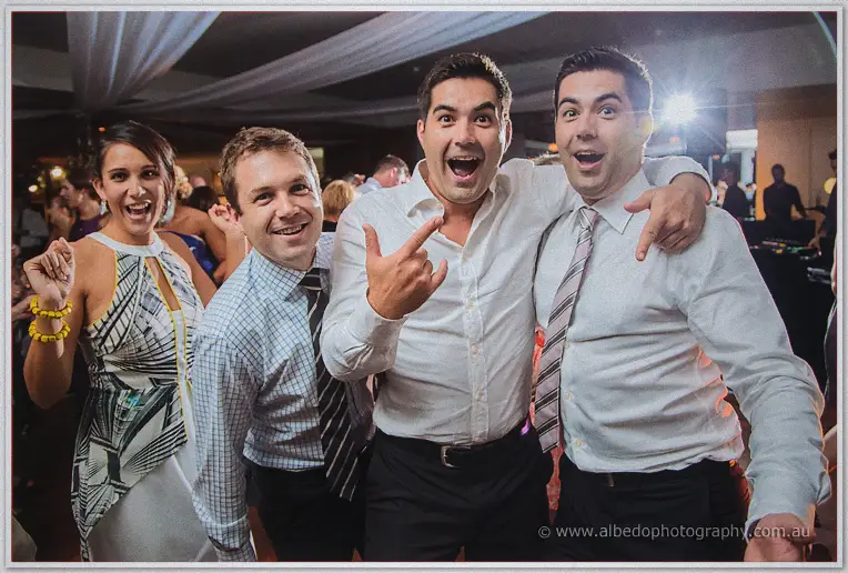 Four party guests pulling wide-eyed rock poses for the camera mid-dance at the crowded reception