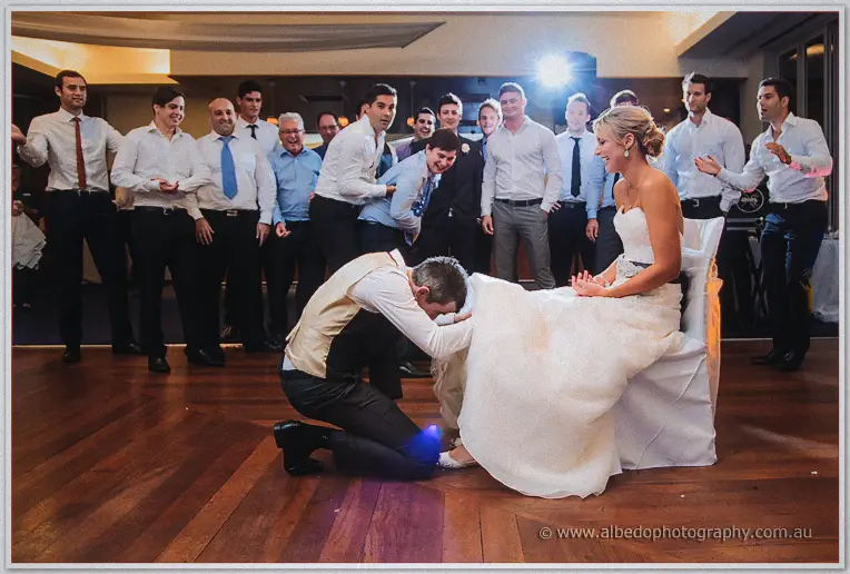 Groomsman hunting for the garter around Lauren's ankle as she sits laughing amid a wall of grinning men